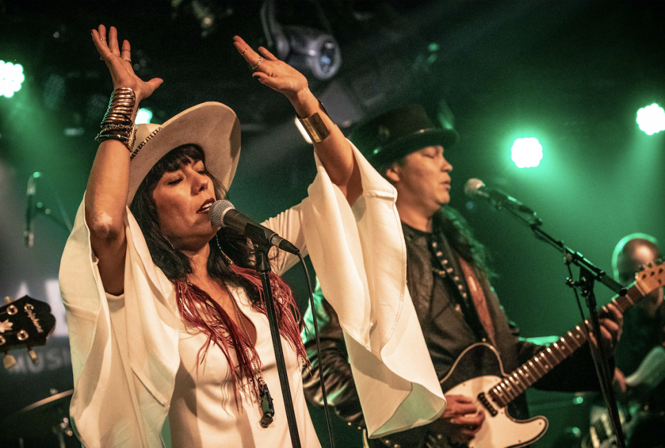 Female singer onstage with hands in the air. Band and green lighting in the background
