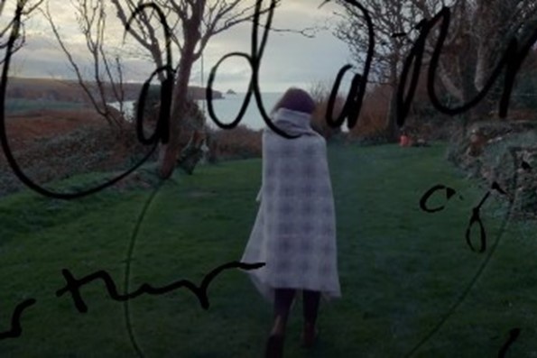 A person wrapped in a blanket with their back to the camera is stood on grass with a house to their right and the sea and mountains to the left. The word 'cadarn' is scribbled across the picture