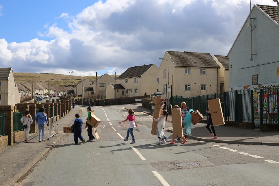 Young people are crossing a road in a Welsh valleys town, carrying cardboard boxes