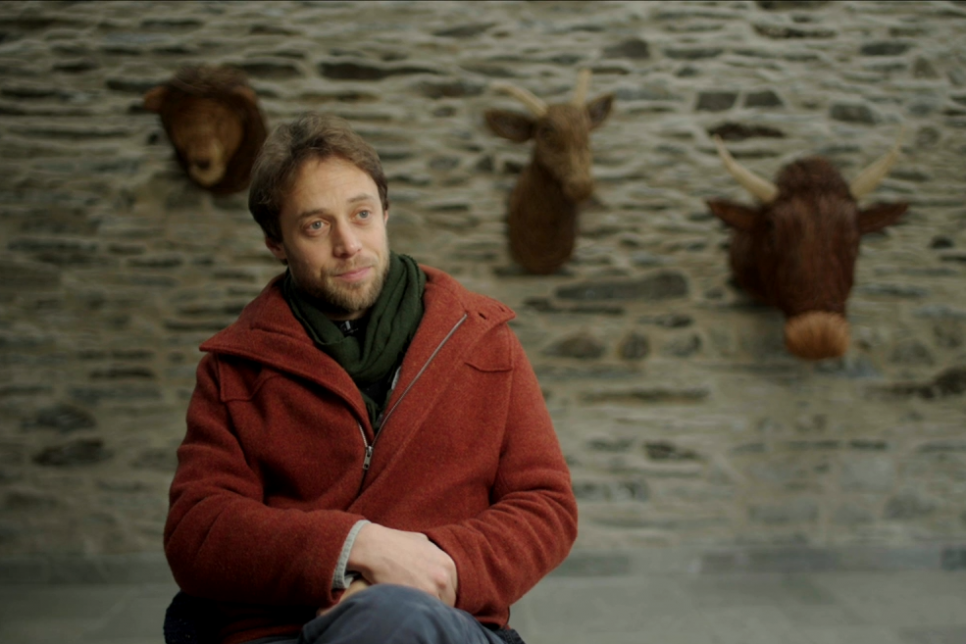 The artist Cynefin sat in a chair in front of a stone wall with stuffed animal art.