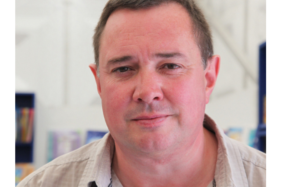 A portrait of the author Jon Gower wearing a light beige shirt. The background is softly blurred emphasizing focus on the person.