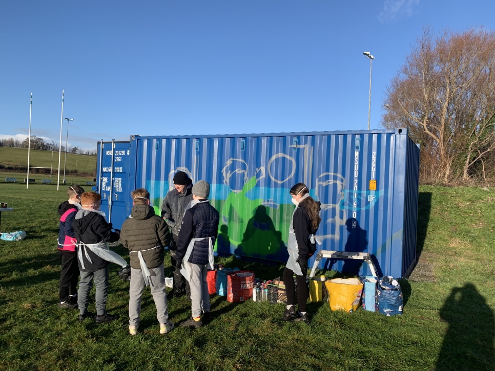 Pobl ifanc a phlant yn paentio mur ar gynhwysydd cludo glas mewn cae