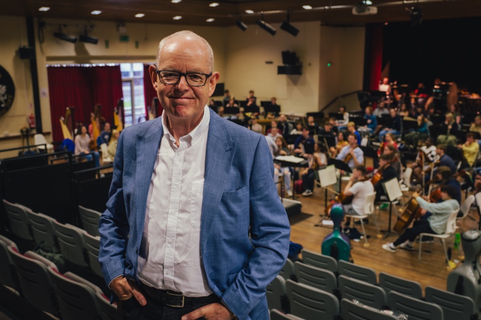 A photograph of a man wearing black framed glasses and a blue blazer in front of a youth orchestra who are rehearsing in the background