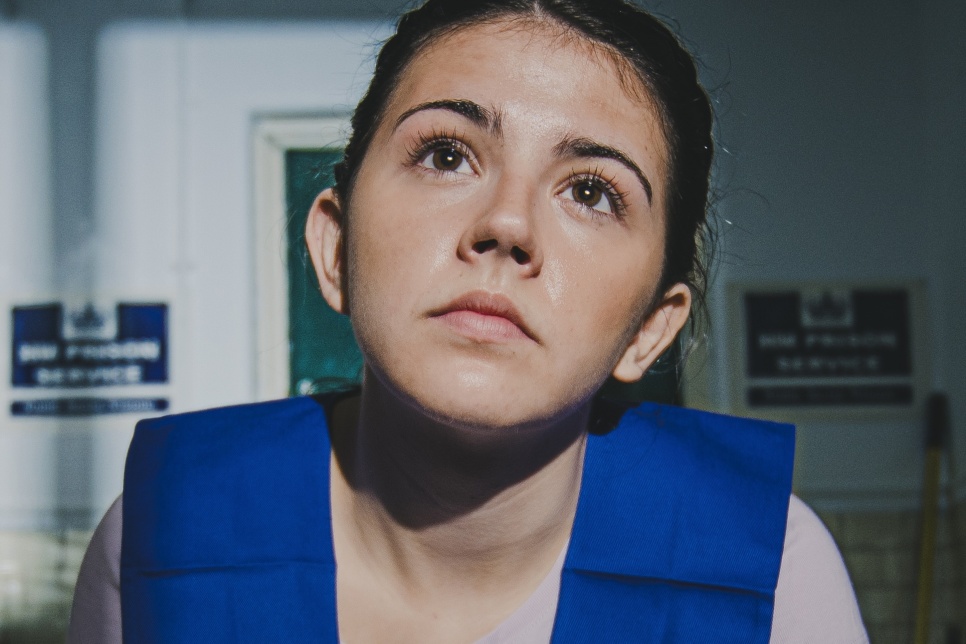 Young woman in prison cell, wearing blue prison bib, looking up wistfully