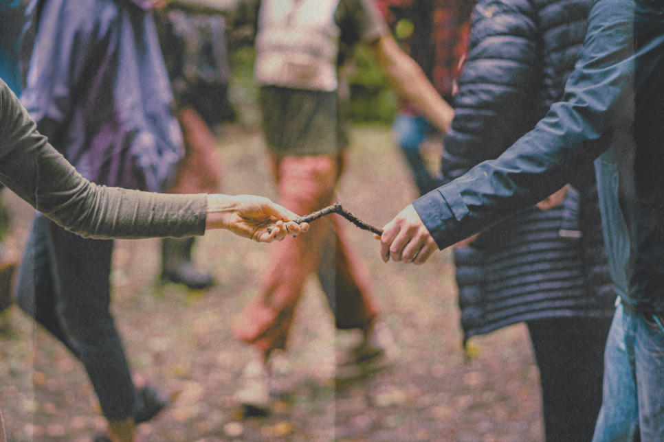 Image of people outdoors. There are two people in the foreground who are holding each end of the same wooden stick / Delwedd o bobl tu allan. Mae dau berson yn y blaendir yn dal yr un brigyn pren ar bob pen