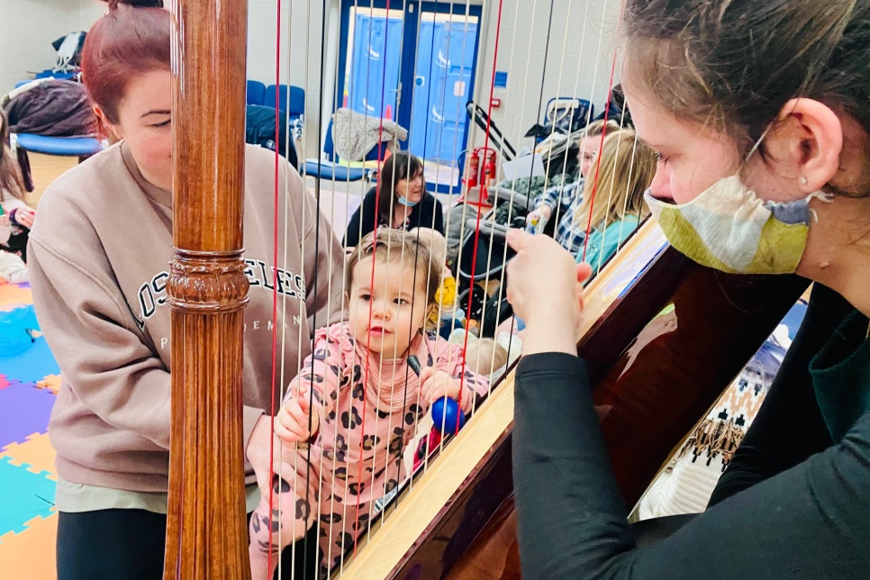 A person playing on a harp while a person and a baby listens