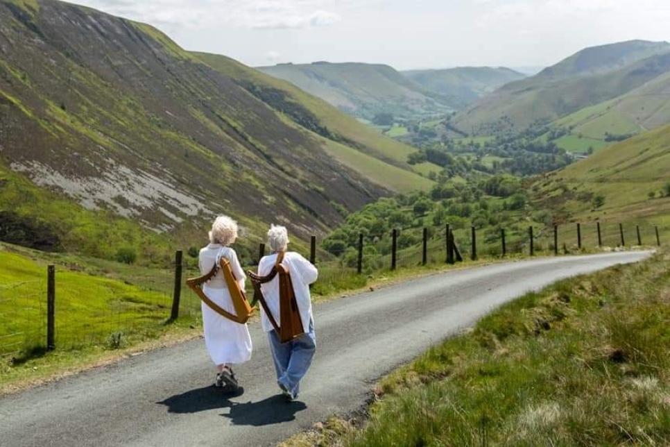Two harpists walking down a mountain road with their harps on their backs
