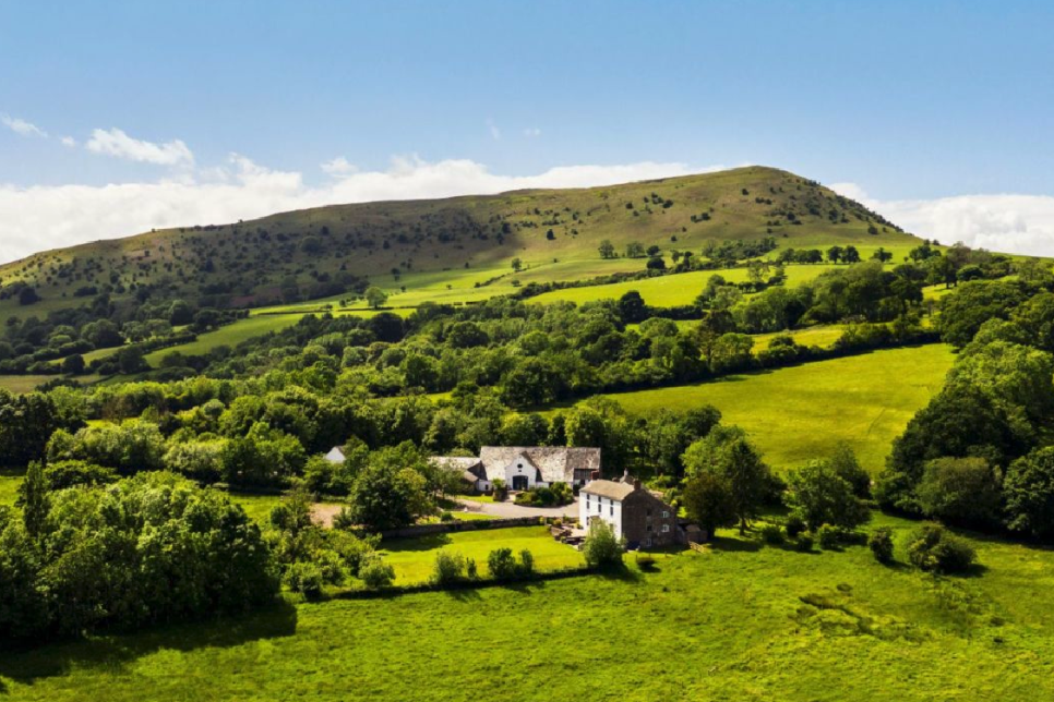 A photograph from a distance of farm buildings and green fields with a hill in the background and blue sky / Ffotograff o bellter o adeiladau fferm a chaeau gwyrdd gyda bryn yn y cefndir ac awyr las 