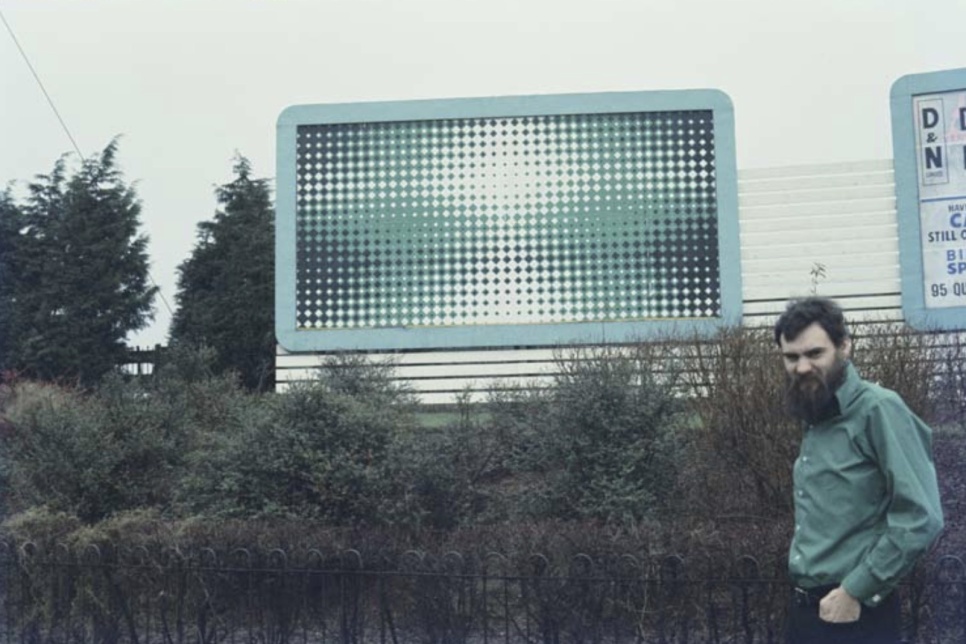 A scan of a photograph taken in 1967. A billboard with a green and black checkered 'op-art' pattern on it is in the centre. To the right in the foreground a man in a green shirt with a beard. 