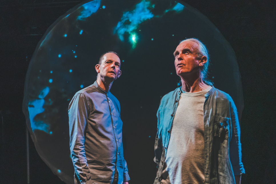 Photo of Daniel Morden and Hugh Lupton performing Stars and their Consolations on stage in front of star images in the background / Ffotograff o Daniel Morden a Hugh Lupton yn perfformio Stars and their Consolations ar y llwyfan o flaen delweddau o sêr yn y cefndir