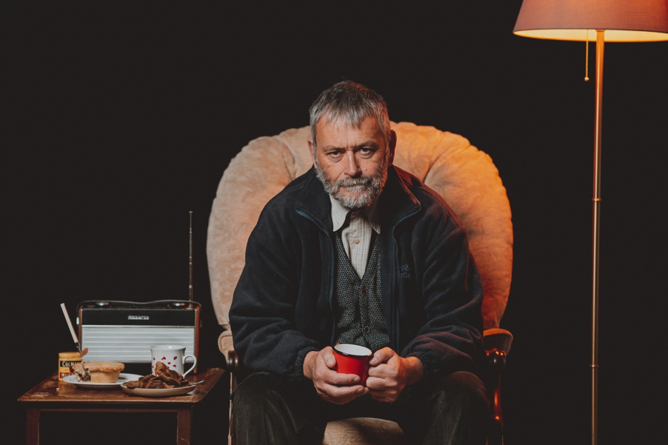 Delwedd o actor ar set lwyfan yn eistedd ac yn dal mwg / Image of an actor on set sitting down and holding a mug 