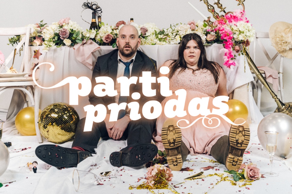 Parti Priodas poster image with Mark Henry and Mared Llywelyn. Both are dresses in formal attire, with Mared in a bridesmaids dress and wellies. Behind them, a wedding top table with flowers, candelabras, and more is in disarray.
