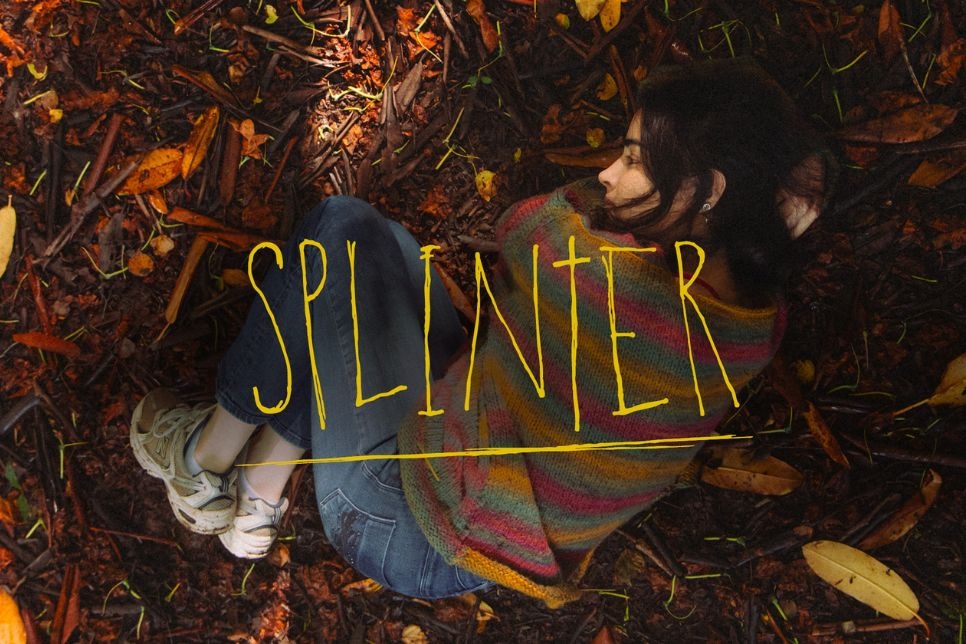 An image of a woman curled up on a floor that is covered with forest litter. The word ‘Splinter’ is written across the middle of the image / Delwedd o fenyw yn gorwedd mewn pelen ar lawr sydd wedi ei orchuddio â dail a gwasarn. Mae’r gair ‘Splinter’ wedi ei ysgrifennu ar draws canol y ddelwedd