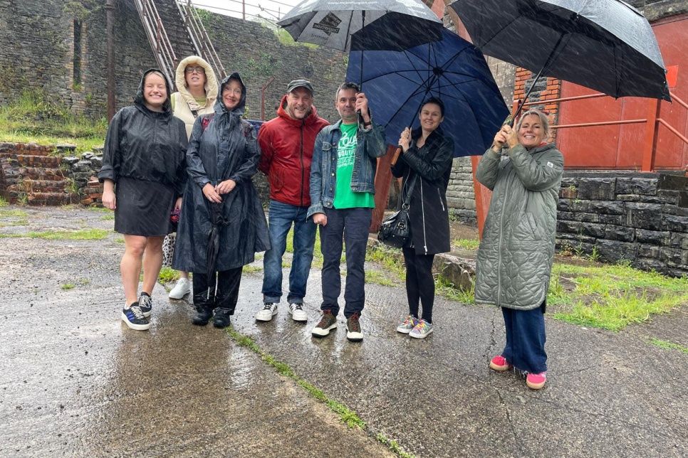 A picture of seven people standing outside on a wet day. Three are holding umbrellas. / Llun o saith o bobl yn sefyll tu allan ar ddiwrnod gwlyb. Mae tri yn dal umbrellas.